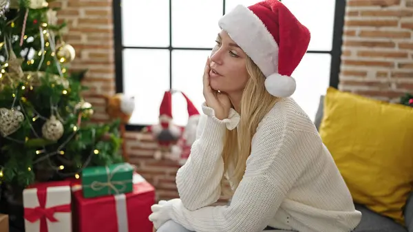 年轻的金发女人戴着圣诞礼帽坐在沙发上 满脸严肃的表情在家里 — 图库照片