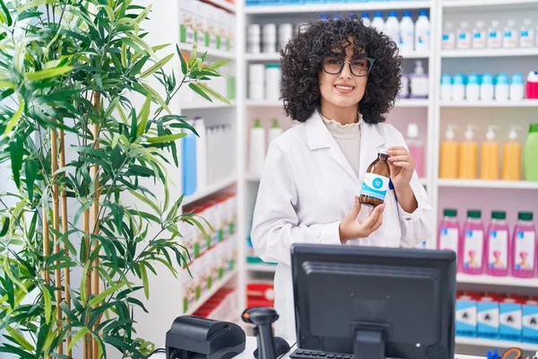 Young middle eastern woman pharmacist smiling confident holding medication bottle at pharmacy
