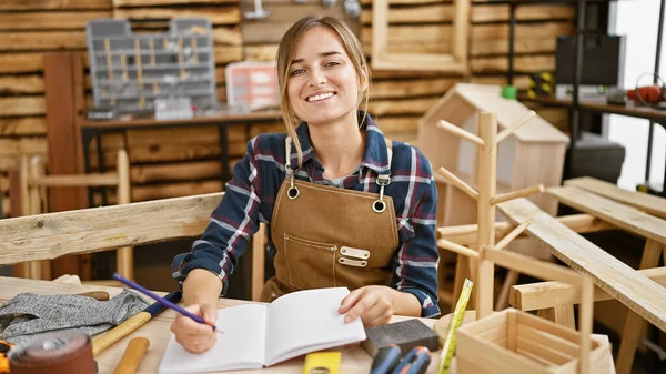 Radiant young blonde woman carpenter, confidently taking notes, exudes joy at her carpentry workshop table