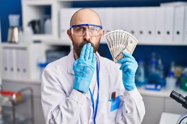 Young Hispanic Man Working Scientist Laboratory Holding Money Covering Mouth — Stock Photo, Image