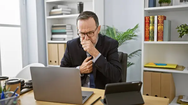 Stressed middle-aged man with grey hair, working hard in business, coughing at his office desk despite illness