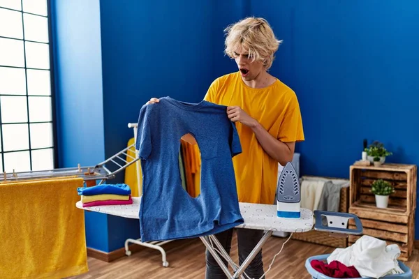 Mladý Blonďatý Muž Drží Tričko Spálenou Dírou Překvapivým Výrazem Prádelně — Stock fotografie