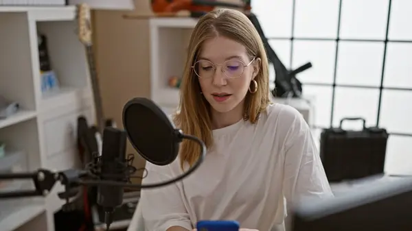 Young blonde woman radio reporter reading smartphone screen speaking on a show at radio studio