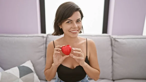Joyful young hispanic woman adoringly holding heart gesture to her chest, sitting relaxed on cozy sofa in living room, flashing confident smile.