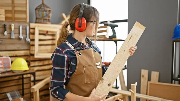 迷人的金发女木匠在木工车间里挥舞着木板 戴着安全的耳机 带着工作节奏 — 图库照片