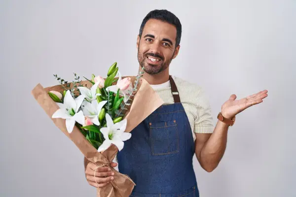 Hispanic man with beard working as florist smiling cheerful presenting and pointing with palm of hand looking at the camera.