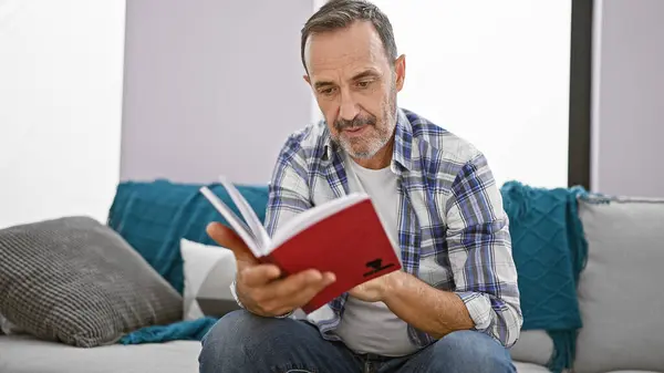 中年男性は灰色の髪で 快適にリビングルームのソファーに座って 自宅で本を読んで吸収しました — ストック写真
