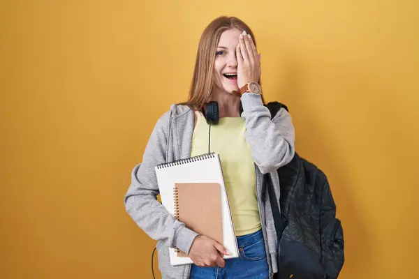Young caucasian woman wearing student backpack and holding books covering one eye with hand, confident smile on face and surprise emotion.