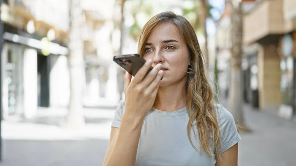 Stunning young blonde woman standing relaxed on city street, engrossed in sending intimate voice message via her smartphone, a portrait of modern technology merging with urban outdoors