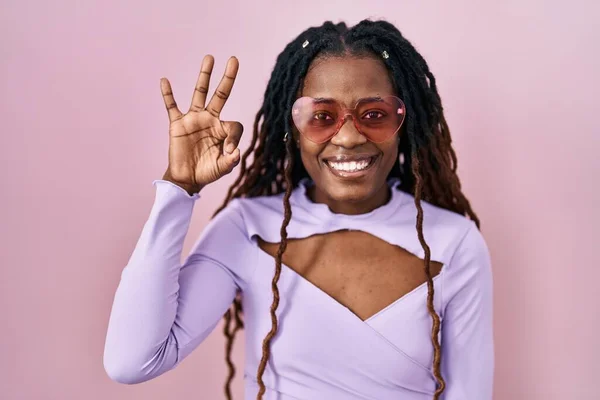 ピンクの背景の上に立っている編まれた髪を持つアフリカの女性は ポジティブに笑顔で手と指でOkサインします 成功した表現 — ストック写真