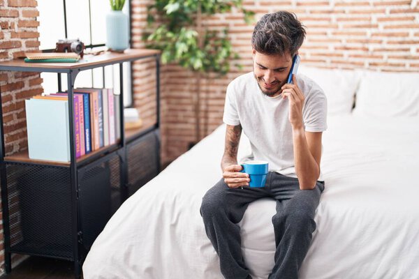Молодой латиноамериканец разговаривает по смартфону, пьет кофе в спальне