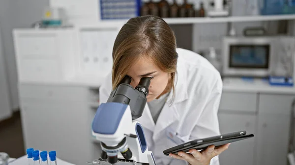 Serious young blonde female scientist engrossed in discoveries, meticulously analyzing samples under a microscope in a hospital laboratory while holding a touchpad.