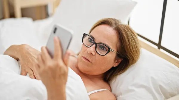 Латиноамериканка Средних Лет Очках Выглядящая Смартфон Лежит Кровати Спальне — стоковое фото