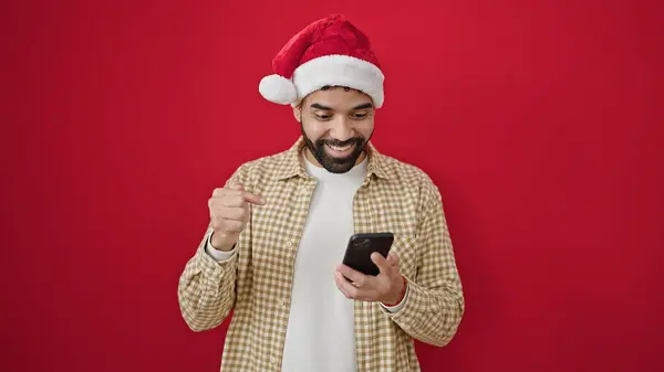 Junger Hispanischer Mann Mit Weihnachtsmütze Zeigt Auf Smartphone Vor Isoliertem — Stockfoto