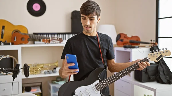 Hispanic young male guitarist, dialing up the electro-tunes, texting while tackling electric guitar in the music studio