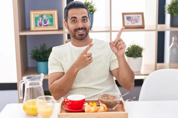Hispanic man with beard eating breakfast smiling and looking at the camera pointing with two hands and fingers to the side.
