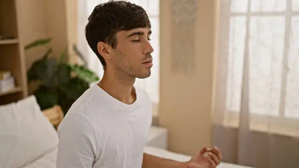 Handsome young hispanic man starts his morning right, concentrating on a calming yoga exercise in his bedroom sitting on the bed under the cosy blanket