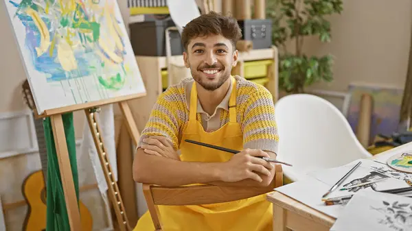 Confident, smiling young arab man artist sitting with arms crossed in his art studio, joyfully embracing his painting hobby