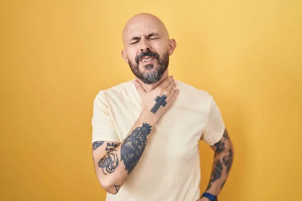 Hispanic man with tattoos standing over yellow background touching painful neck, sore throat for flu, clod and infection