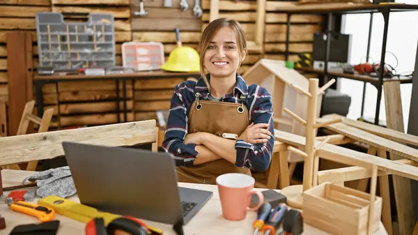 Smiling young blonde woman carpenter, arms crossed, joyfully navigating carpentry business on laptop amidst sawdust and timber in indoor workshop