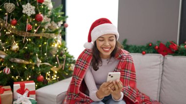Genç ve güzel İspanyol kadın akıllı telefon kullanıyor. Yılbaşı ağacının yanında oturuyor.
