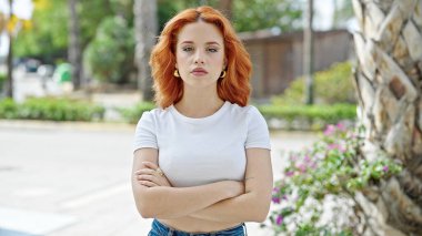 Genç kızıl saçlı kadın parkta sakin bir ifadeyle ve kollarını kavuşturarak duruyor.