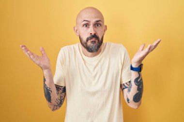 Sarı arka planda dikilen İspanyol dövmeli, elleri ve kolları havada, kafası karışmış bir adam. şüphe kavramı. 