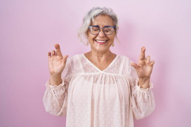 Middle age woman with grey hair standing over pink background gesturing finger crossed smiling with hope and eyes closed. luck and superstitious concept. 