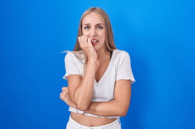 Young caucasian woman standing over blue background looking stressed and nervous with hands on mouth biting nails. anxiety problem. 