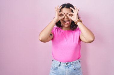 Young hispanic woman standing over pink background doing ok gesture like binoculars sticking tongue out, eyes looking through fingers. crazy expression. 