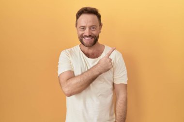 Orta yaşlı, sakallı, sarı arka planda neşeli, yüzünde bir gülümseme, mutlu ve doğal bir ifadeyle elini ve parmağını kaldırmış bir adam. 