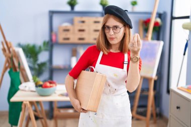 Sanat stüdyosundaki genç kızıl saçlı kadın elinde sanat çantası tutarak italyan jestleri yapıyor. 