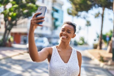 Afro-Amerikalı kadın gülümsüyor. Sokakta akıllı telefonun yanında özçekim yapıyor.