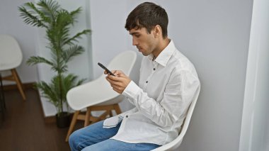 Ciddi, genç İspanyol bir adam, başarılı bir profesyonel, sandalyede rahat bir şekilde oturuyor, akıllı telefonuyla mesaj atıyor, geniş bir bekleme odasında önemli bir iş görüşmesi bekliyor.