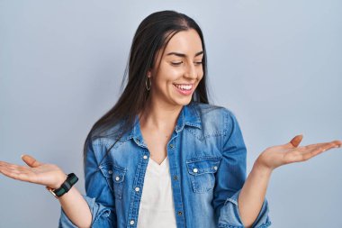 Hispanic woman standing over blue background smiling showing both hands open palms, presenting and advertising comparison and balance 