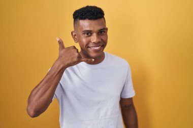 Young hispanic man standing over yellow background smiling doing phone gesture with hand and fingers like talking on the telephone. communicating concepts. 