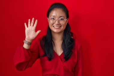 Asyalı genç bir kadın kırmızı arka planda duruyor. Beş numarayı işaret ediyor ve gülümsüyor. Kendinden emin ve mutlu.. 