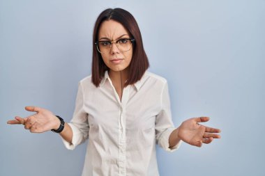 Young hispanic woman standing over white background clueless and confused with open arms, no idea concept. 