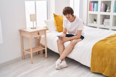 Genç sarışın adam yatakta akıllı telefon kullanıyor.