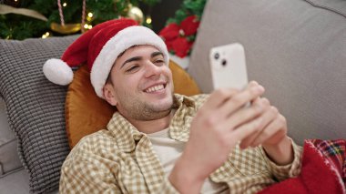 Evdeki Noel ağacının yanındaki koltukta akıllı telefon kullanan İspanyol genç adam.