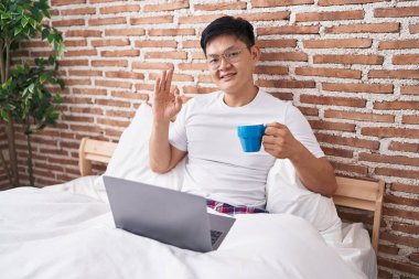 Genç Asyalı adam yatakta kahve içiyor parmaklarıyla imza atıyor, gülümsüyor ve mükemmel bir sembol oluşturuyor. 