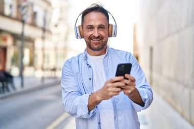 Genç, beyaz bir adam gülerek sokakta müzik dinliyor.