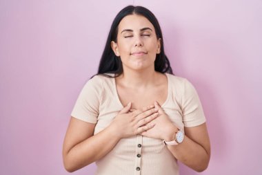 Pembe arka planda duran genç İspanyol kadın elleri göğsünde, gözleri kapalı ve yüzünde minnet dolu bir jest ile gülümsüyor. sağlık kavramı. 