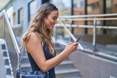 Genç, güzel, İspanyol bir kadın sokakta akıllı telefon kullanarak gülümsüyor.