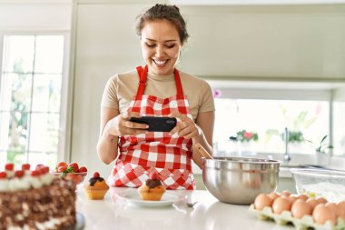 Genç, güzel, kendine güvenen İspanyol bir kadın. Akıllı telefondan kek yapıp mutfakta fotoğraf çekiyor.
