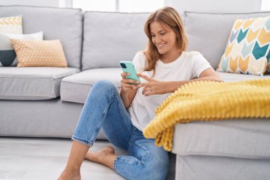 Young blonde girl using smartphone sitting on floor at home