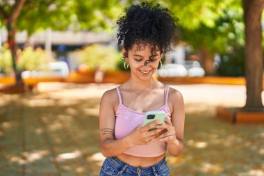 Genç İspanyol kadın parkta akıllı telefon kullanarak gülümsüyor.