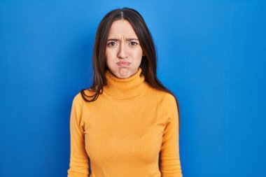 Genç esmer kadın mavi arka planda duruyor komik yüzüyle yanaklarını şişiriyor. Havayla şişmiş ağız, çılgın bir ifade.. 