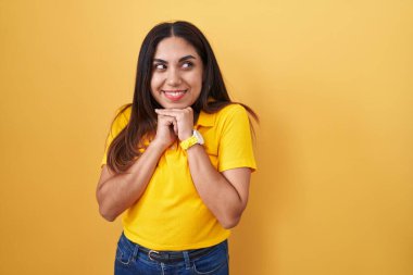 Sarı arka planda dikilen genç Arap kadın. Gergin ve heyecanlı. Elleri çenesinde yan tarafa bakıyor. 