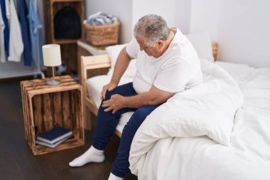 Middle age grey-haired man suffering for knee injury sitting on bed at bedroom